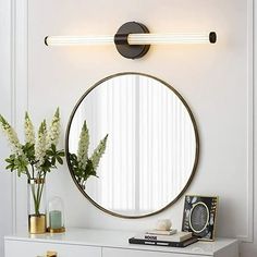 a round mirror mounted on the wall above a white dresser with flowers in vases