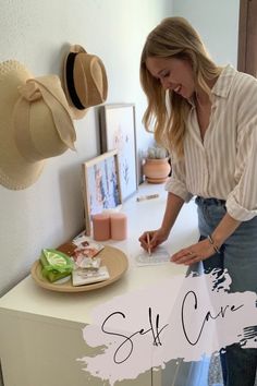 a woman standing in front of a white counter with hats on it and writing self care