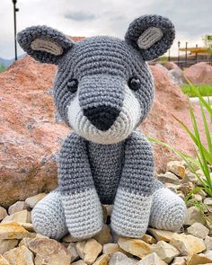 a crocheted gray and white bear sitting on top of rocks next to grass