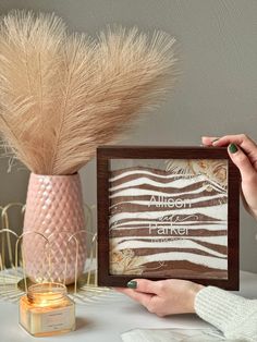 a woman holding up a framed photo next to a vase with some dry grass in it