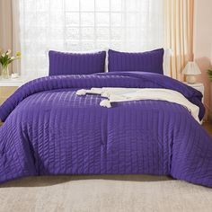 a bed with purple comforter and pillows in front of a white curtained window