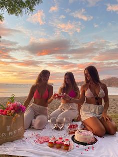 three women in bikinis sitting on a blanket at the beach with cupcakes