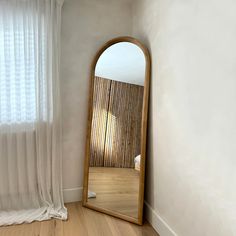 a large mirror sitting on top of a wooden floor next to a white curtained window