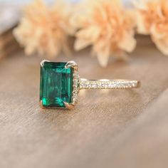 an emerald and diamond ring sitting on top of a piece of wood with flowers in the background