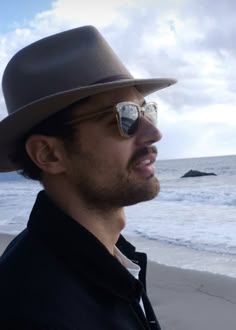 a man wearing sunglasses and a hat standing on the beach next to the ocean with a no swimming sign in front of him