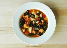 a white bowl filled with soup and vegetables on top of a wooden table next to a spoon