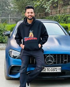 a man standing in front of a blue car wearing a black sweatshirt and sweatpants