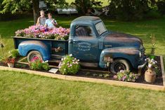 two people are sitting in the back of an old truck with flowers growing out of it