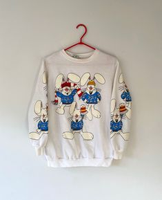 Rare! this colorful novelty pullover sweater from the 90s features an all over print of happy cartoon bunnies. there are three bunnies on the front of the sweater and two on each of the sleeves. the bunnies are each wearing blue sweaters with white bunnies on them, and some of the bunnies are also wearing toques and scarves, and the white paint of the bunnies are slightly puffy. the sweater has a white (slightly sheer) body, long sleeves and a crew neckline. the fabric is a soft cotton polyester Playful Crew Neck Sweater With Cartoon Print, Multicolor Crew Neck Sweatshirt With Cartoon Print, Fun Cartoon Print Sweatshirt, Playful Crew Neck Sweater With Graphic Print, Cartoon Bunnies, Cartoon Animals Cute, Three Bunnies, White Bunnies, Happy Cartoon