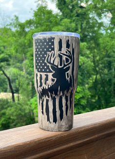 a tumbler cup sitting on top of a wooden ledge with trees in the background