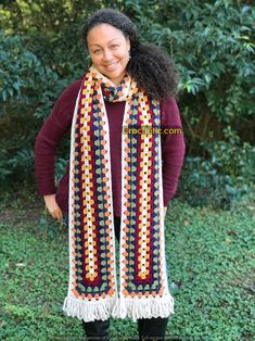 a woman wearing a multicolored crocheted scarf standing in front of some bushes