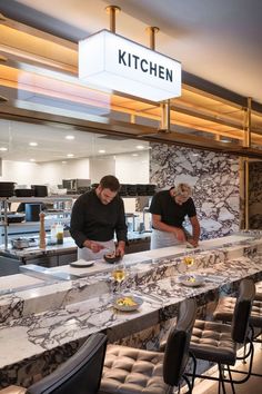 two men preparing food in a kitchen with marble counter tops and bar stools around them