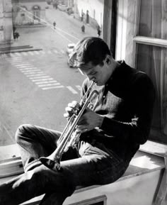 a man sitting on a window sill holding a trumpet in his right hand and looking down at the ground