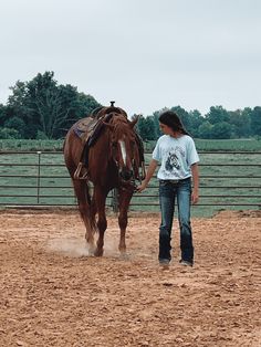 a woman standing next to a brown horse on top of a dirt covered field in front of a fence