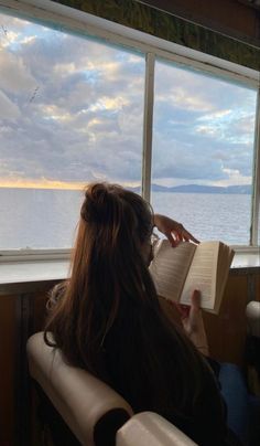 a woman reading a book while looking out the window at the water and clouds outside