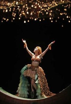 a woman in a dress is standing on a stage with her arms outstretched and lights hanging from the ceiling