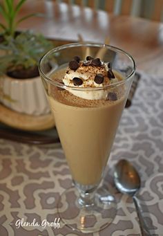 a dessert in a glass on top of a table next to a spoon and potted plant