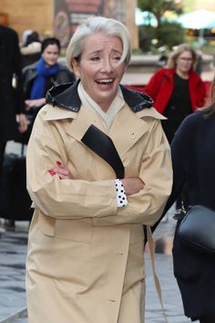 an older woman in a trench coat smiles as she walks down the street with her arms crossed