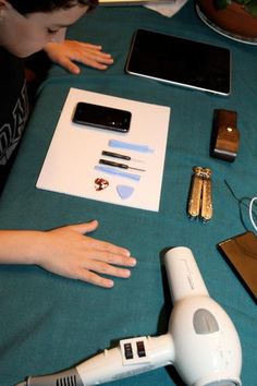 a young boy sitting at a table with electronics and other items on it, including a hair dryer