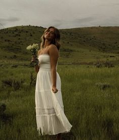 a woman in a white dress is standing in the grass and holding a bunch of flowers