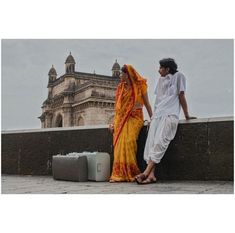 a man and woman standing next to each other near a wall with an old building in the background