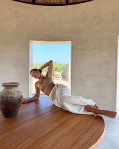 a nude woman laying on top of a wooden table next to a vase and window