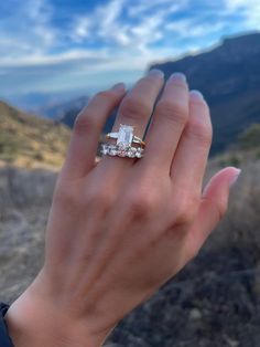 a woman's hand holding an engagement ring in front of a mountain range with blue sky and clouds