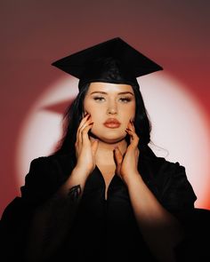 a woman wearing a black graduation gown and holding her hands to her face while standing in front of a red background