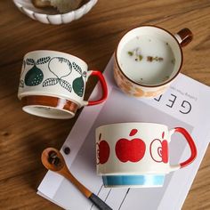 two coffee mugs sitting on top of a wooden table next to a book and spoon