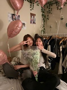 two women sitting on a bed with balloons and flowers in front of them, posing for the camera