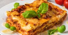 a white plate topped with lasagna covered in cheese and basil next to tomatoes