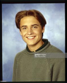 a young man in a green sweater smiling at the camera with a blue sky behind him