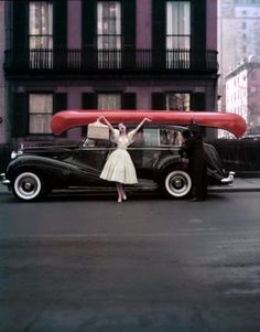 a woman standing next to a black car with a red surfboard on it's roof