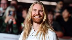 a man with long blonde hair and beard smiles at the camera as he stands in front of a crowd