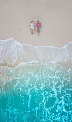 two people laying on the beach with their arms around each other and looking at the water