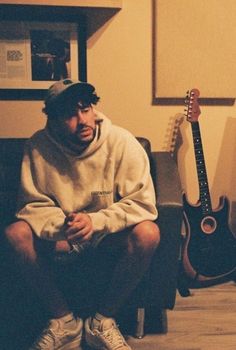 a man sitting on the floor next to an electric guitar and wearing a hoodie