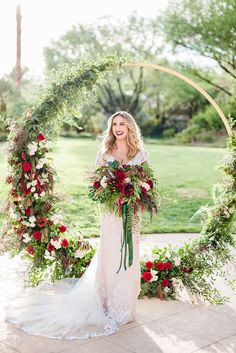 a woman standing in front of an arch with flowers and greenery on it, holding a bouquet