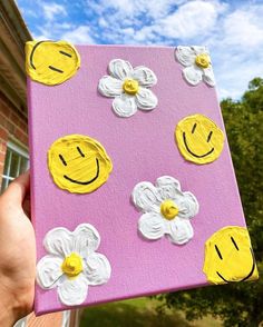 a hand holding up a painted canvas with flowers and smiley faces on it in front of a house