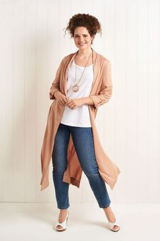 a woman standing in front of a white wall wearing jeans and a tan cardigan