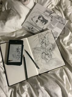 an open book on top of a bed next to a pen and drawing pad with drawings