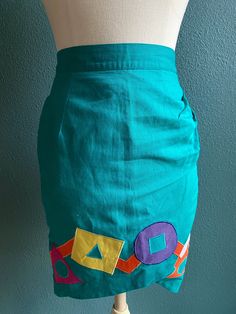 Vintage Skirt in Bright Teal Green with multi color geometric graphic shapes appliquéd on  -skirt is tulip shaped  -zips and buttons in the back  -waist has elastic in part of band  -Made by Deborah Lunt Boutique  Measurements laying flat: waist 12.5 inches / stretched 14 inches  Length 22.5" Blue Fitted Patchwork Mini Skirt, Fitted Blue Patchwork Mini Skirt, Blue Fitted Cotton Pencil Skirt, Blue Fitted Mini Skirt With Patchwork, Fitted Blue Mini Skirt With Patchwork, Spring Retro Fitted Pencil Skirt, Retro Fitted Pencil Skirt For Spring, Retro Fitted Mini Skirt With Pockets, Fitted Patchwork Mini Skirt For Spring