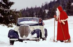 a man dressed as santa claus standing next to an old fashioned car in the snow