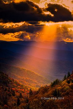 the sun shines brightly through clouds over mountains and trees in fall colors, as seen from an overlook