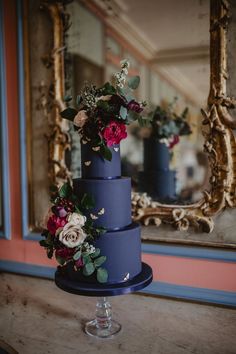 a three tiered blue cake with flowers on top sits in front of a mirror