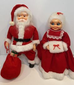 two santa claus dolls sitting next to each other on a white surface with a red bag in front of them