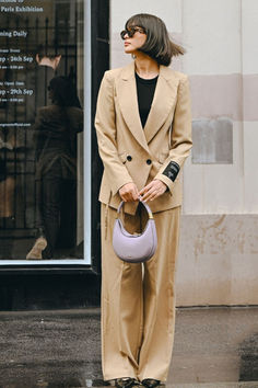 a woman standing in front of a building holding a purple purse and wearing a tan suit