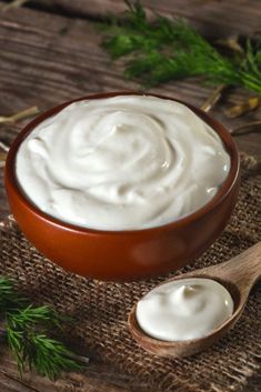 a bowl of yogurt next to a spoon on a wooden table with green sprigs