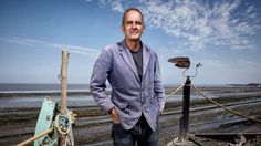 a man standing next to a boat on the beach with his hands in his pockets