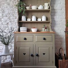 an old fashioned hutch with dishes and cups on the top, in front of a brick wall