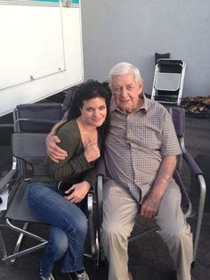 an older man sitting next to a woman in a chair on the ground with her arm around him